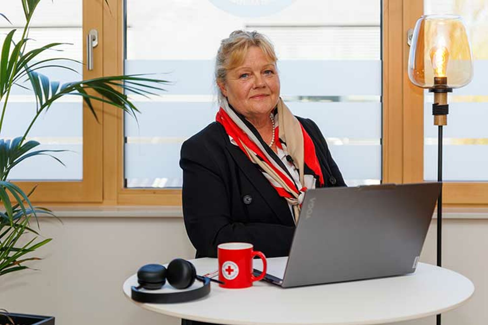 Pflegeberaterin Martina Ohrmann von der DRK ambulante Pflege Mittelhessen gGmbH sitzt mit Laptop und roter DRK-Tasse an einem Tisch und berät online.